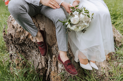 cropped photo of the bride and groom sitting on a tree stump in the forest in elegant attire. Bouquet of flowers from peonies, red shoes of the groom