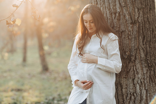 A pregnant woman in a white dress on a dark background poses in the park. Happy and carefree pregnancy. Autumn park. Happy woman enjoying her pregnancy.