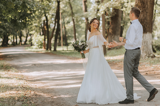 A young couple of newlyweds are walking in a summer park. The bride in a beautiful white dress with a wreath of fresh flowers on her head, the groom in a white shirt