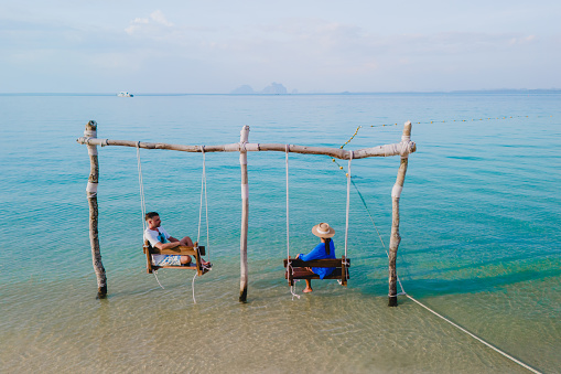 a couple of men and a woman on a swing at the beach of Koh Muk tropical island , and a turqouse colored ocean in Koh Mook Trang Thailand, a couple on a swing in the ocean