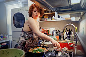Morning meal preparation. Mature ginger woman in nightgown, cooking omelette with vegetables