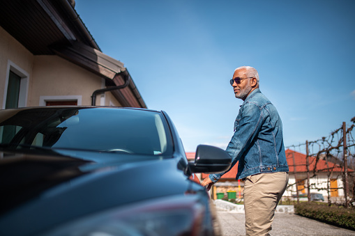 Elderly Hispanic male in casual attire preparing to enter a car, outdoors with residential background.