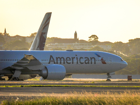 An American Airlines Boeing B777-323(ER) plane, registration N720AN, parked at Sydney Kingsford-Smith Airport.  In the background is the vertical stabiliser of a Qatar Airways Airbus A380-861 plane, registration A7-APE, taxiing after arrival from Doha as flight QR908. This image was taken near General Holmes Drive, Mascot on a hot and sunny day near sunset on 9 March 2024.