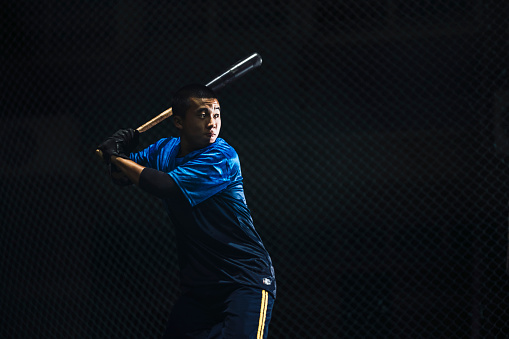 This is a photo of a baseball player practicing hitting skills with focus and passion during a night-time baseball training session. The motion of him swinging the bat and hitting the baseball is captured in the photo.