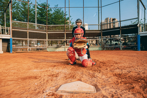 The catcher standing behind home plate gazes through the slits of his mask at the camera. He is responsible for catching the pitches thrown by his teammates and contributing to the team's defense.
