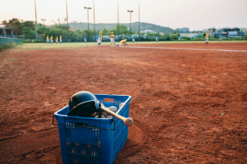 Baseball field with baseball equipment including baskets filled with baseballs and bats with players practicing in the background