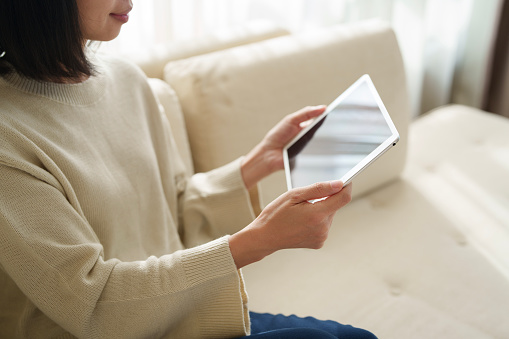 In the close-up shot, an Asian woman is sitting on a sofa, her hand is seen using a digital tablet while working from home, with sunlight gently shining on her. This illustrates the blend of lifestyle, technology, and relaxation.