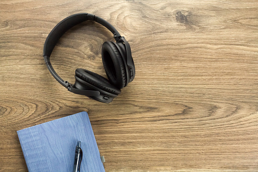 Overhead view of wireless headphones and notebook with pen on a wooden table. Top down view of black over ear headphones on a wooden table