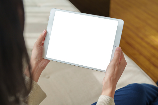 In the close-up shot, an Asian woman is sitting on a sofa, her hand is seen using a digital tablet while working from home, with sunlight gently shining on her. This illustrates the blend of lifestyle, technology, and relaxation.