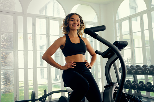Happy young multiracial woman wearing sports clothing on exercise bike at the gym