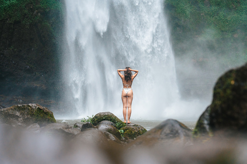 Serene woman standing near the refreshing powerful  waterfall on Bali island