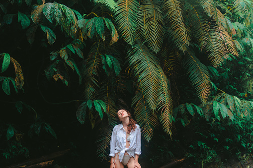 Serene woman in white shirt standing on the background of lush tropical foliage and enjoying serenity