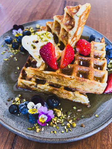 Vertical high angle closeup photo of two freshly baked waffles with strawberries, blueberries, mascarpone, pistachio pieces, maple syrup and viola flowers on a grey ceramic plate on a wooden table in a restaurant.