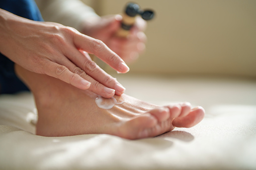In the cropped image, a mid-adult Asian woman is shown applying moisturizing cream to her dry skin on her feet. This nurturing gesture reflects her self-care routine and the effort she puts into maintaining her skin's health and hydration.