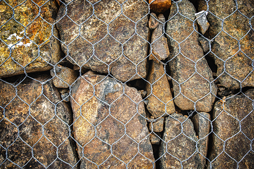 Close-up of a gabion made of brown, rusty, gray stones behind a metal mesh. Fencing, protection, construction.