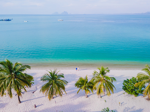 Drone view at a couple walking on the white sandy tropical beach of Koh Muk with palm trees soft white sand, and a turqouse colored ocean in the evening in Koh Mook Trang Thailand