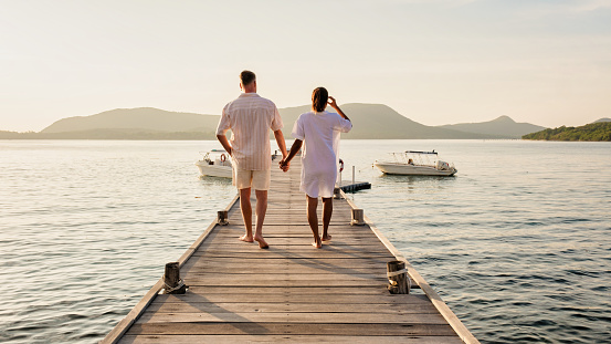 A man and a woman standing on a dock by a lake, holding hands. They are wearing shorts and looking out at the horizon, happy and relaxed during their leisurely travel in Thailand, a couple wooden pier