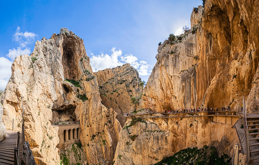 People walking on the Caminito del Rey, a long walkway hanging over 100 metres up on a sheer cliff face in a narrow gorge that runs through cliffs, canyons, and a large valley (8 shots stitched)