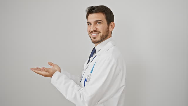 Cheerful young hispanic guy, a bearded doctor wearing a stethoscope, happily gesturing to copy space aside, presenting an excited ad on an isolated white wall, smiles encouraging welcome.