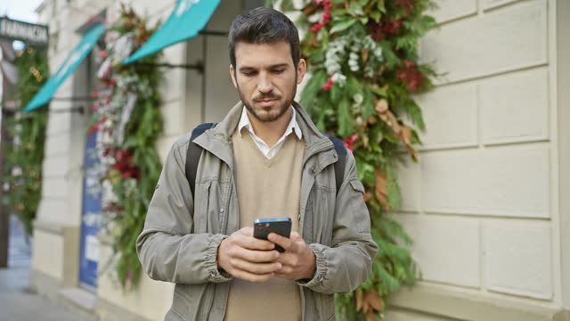A young hispanic man with a beard checks his phone on a decorated city street.