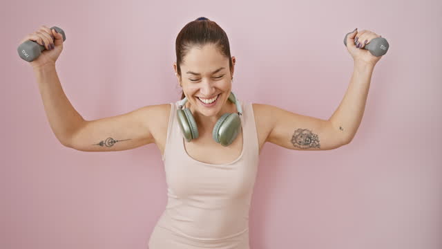 Laughing hard out loud, young woman in sportswear enjoys fun joke while doing weights. blue-eyed beauty in headphones, isolated over pink background, radiates joyful confidence.