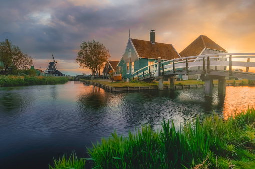 At sunrise, the iconic Zaanse Schans village near Amsterdam, Netherlands, bathes in golden light, casting a sun flare over the traditional wooden green house perched atop the bridge against river and green meadow.