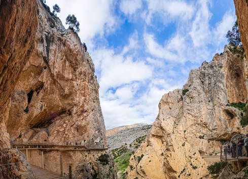 People walking on the Caminito del Rey, a long walkway hanging over 100 metres up on a sheer cliff face in a narrow gorge that runs through cliffs, canyons, and a large valley (6 shots stitched)
