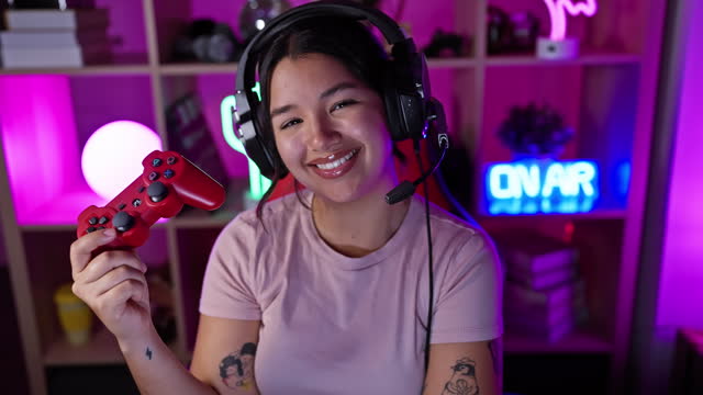 Young hispanic woman smiling with headphones and controller in a luminous gaming room at night