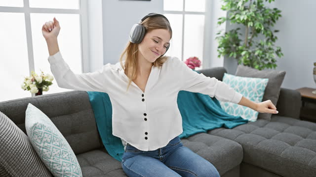 A joyful young woman listens to music with headphones in a cozy living room, expressing happiness.