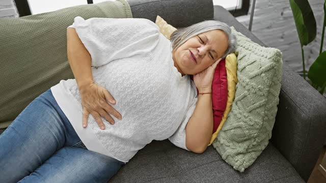 A distressed senior woman with gray hair clutches her stomach lying on a couch indoors, signaling pain or discomfort.