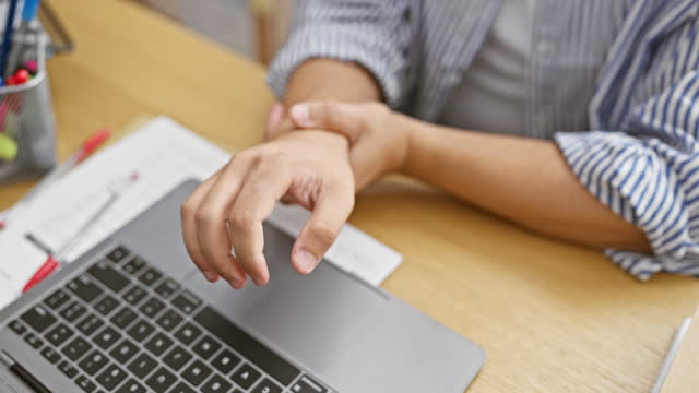 An adult man in an office holding his wrist in pain near a laptop and paperwork, suggesting a possible strain or injury.