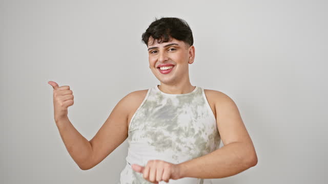Handsome young man confidently presenting in sleeveless t-shirt, smiling while pointing backwards with thumbs up, standing isolated against white background