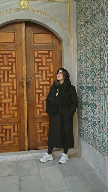 A woman in a hijab leans against an intricately carved wooden door framed by ornate tilework, evoking ottoman istanbul.