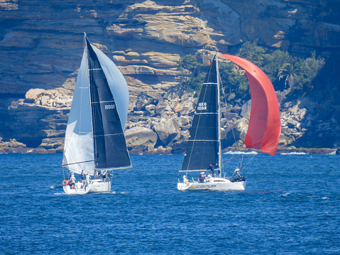 Two yachts under spinnaker sailing on Sydney Harbour.  In the background is the sandstone cliff face of South Head. This image was taken from Manly on a hot and sunny afternoon on 9 March 2024.
