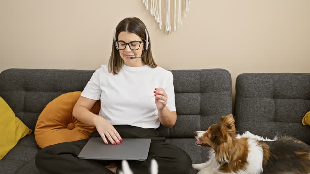 A focused young woman works from home, wearing a headset, with a playful dog on the couch beside her.