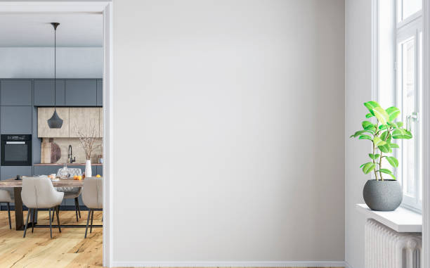 A front view of a modern home interior: a white blank wall with copy space, a window , and a potted plant Ficus on a window sill, a radiator heater under the sill in the foreground.  In the background there is a full dining table (oranges, coffee cups, pastries, books, a vase with flowering branches) and chairs in front of a of a high modern light grey and black glass kitchen cabinets on the hardwood parquet floor.  A built-in coffee machine, 2 ovens, kitchen robot, dishes, plates, pans and other appliances on the countertop in front of the hardwood backsplash. 3D rendered image.