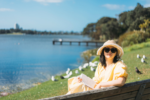 Young Asian woman sitting at the bench near the lake, looking at camera and  holding a book. There are birds, lake and sky in the background.