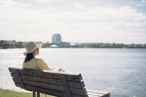 Young Asian woman sitting at a bench by the lake, gazing into distance