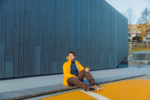 Portrait of a male in yellow jacket sitting in modern yellow skate park admiring beautiful sunny day in Scandinavian town in Norway