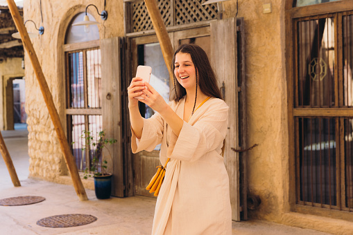 Beautiful female in traditional Arabic dress walking the old streets taking photos of authentic Persian buildings by her smartphone at Al Bastakya district of Dubai city, United Arab Emirates