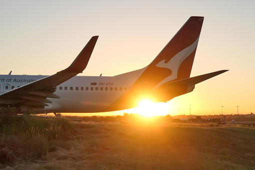 The vertical stabiliser and fuselage of a Qantas Boeing B737-838 plane, registration VH-VYH, taxiing to the third runway in preparation for departure as flight QF550 to Brisbane.  This image faces the setting sun and was taken near General Holmes Drive, Mascot on a hot and sunny day at sunset on 9 March 2024.