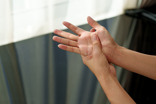 Cropped image of an Asian woman massaging her arthritic hand and wrist, with sunlight shining on her hands, indicating she is experiencing pain and rheumatism