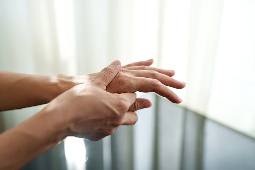 Cropped image of an Asian woman massaging her arthritic hand and wrist, with sunlight shining on her hands, indicating she is experiencing pain and rheumatism