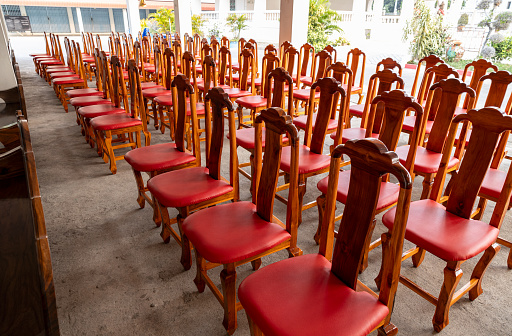 View of wooden chairs and red cushions arranged in rows on the concrete floor outside under a building near an iron gate inside the grounds of a Thai temple.