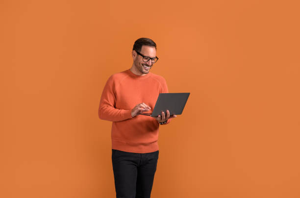 istock Smiling male manager checking e-mails over laptop while standing confidently on orange background 2073371947