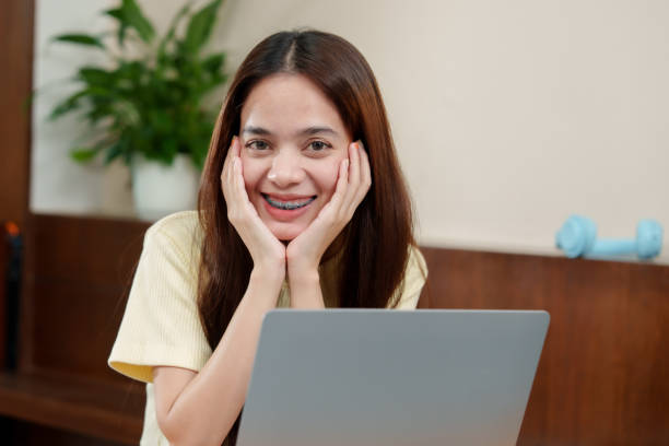 istock Content young Asian woman with braces using a laptop, hands on face, expressing joy in a homely atmosphere. Smiling person with straight hair and braces looks at screen, delight evident 2073381358