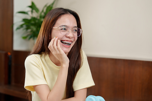Lady with glasses, laughing, hand to mouth, braces visible, bright indoor lighting, joyful ambiance. Exultant in spectacles, highlights dental braces with chuckle, warmly lit room, embodying joviality