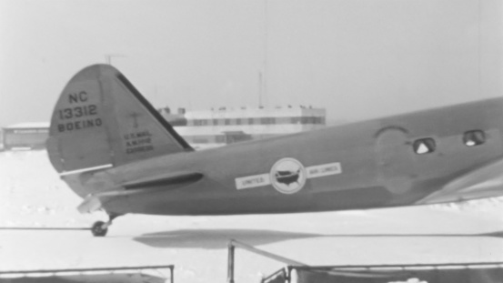 Evocative black and white image of a 1930s propeller airliner. The photo shows the tail and passenger door, with the aircraft parked on a snowy airport. Boeing 247 of United Air Lines