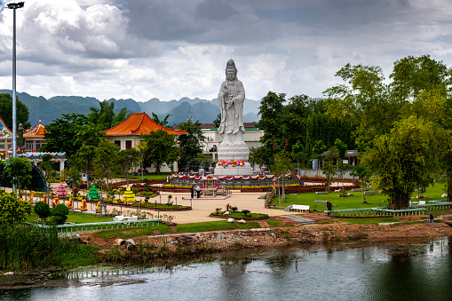 Along the River Kwai towers a Buddhist statue and several traditional style buildings that stand out against the green trees and mountain scenery in the background.