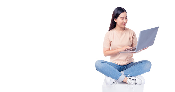 Happy young asian woman sitting on white chair with legs crossed and using laptop computer on light pink background copy space Full body young smiling happy female wearing t shirt and jeans lifestyle